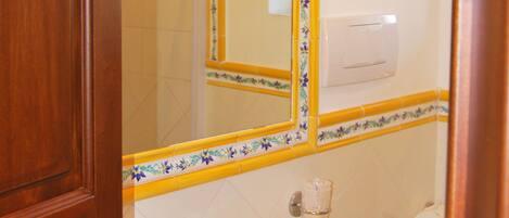 A bathroom with a wooden door, a white sink, a mirror with a floral border, a light fixture, and a toilet.