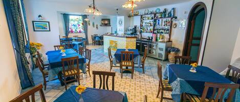 Un comedor con varias mesas y sillas, una zona de bar con estantes llenos de botellas, y una cocina al fondo.