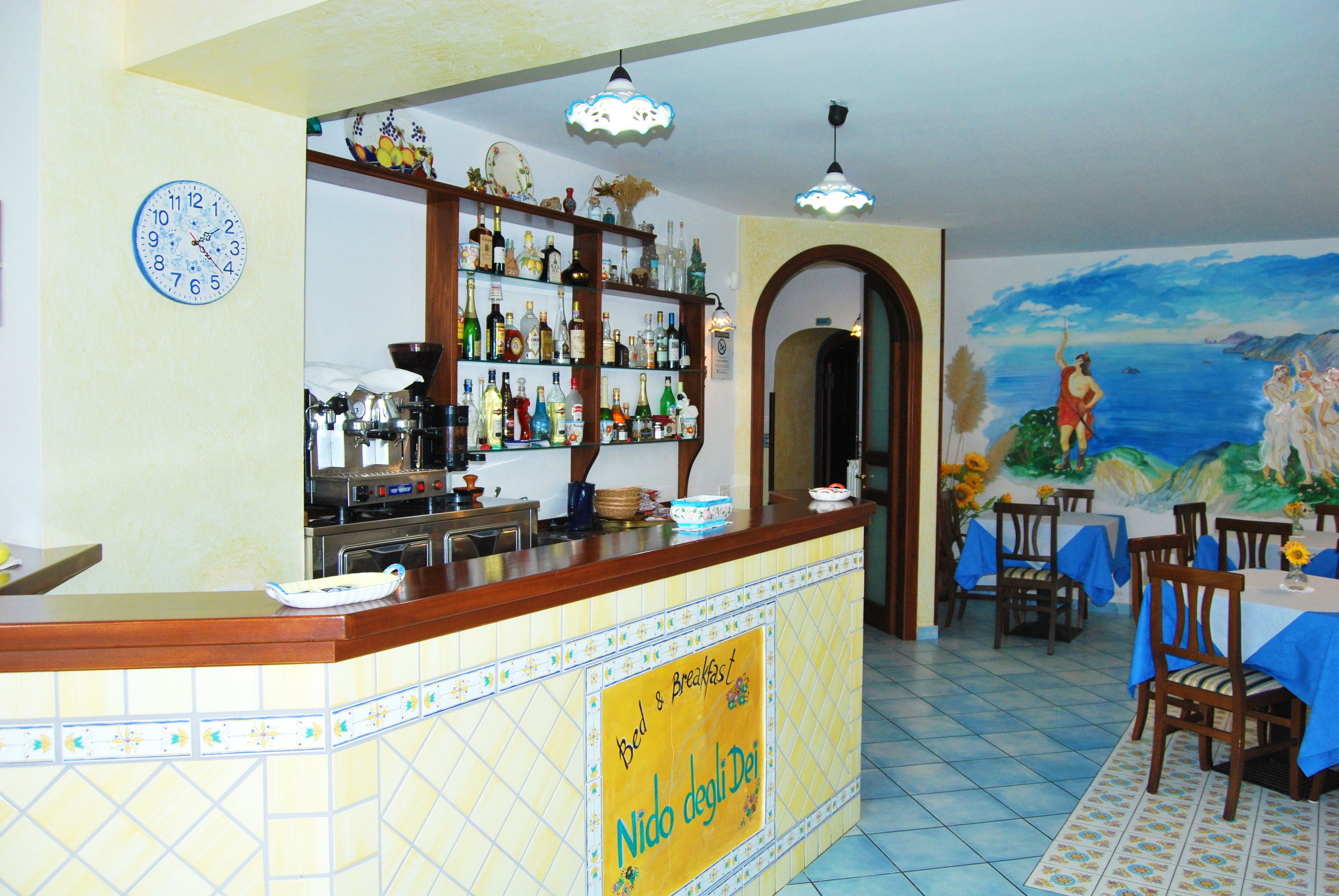 Un restaurante con suelo de baldosas, una barra con un letrero que dice "Bed & Breakfast Nido degli Dei" y un mural de una escena de playa.