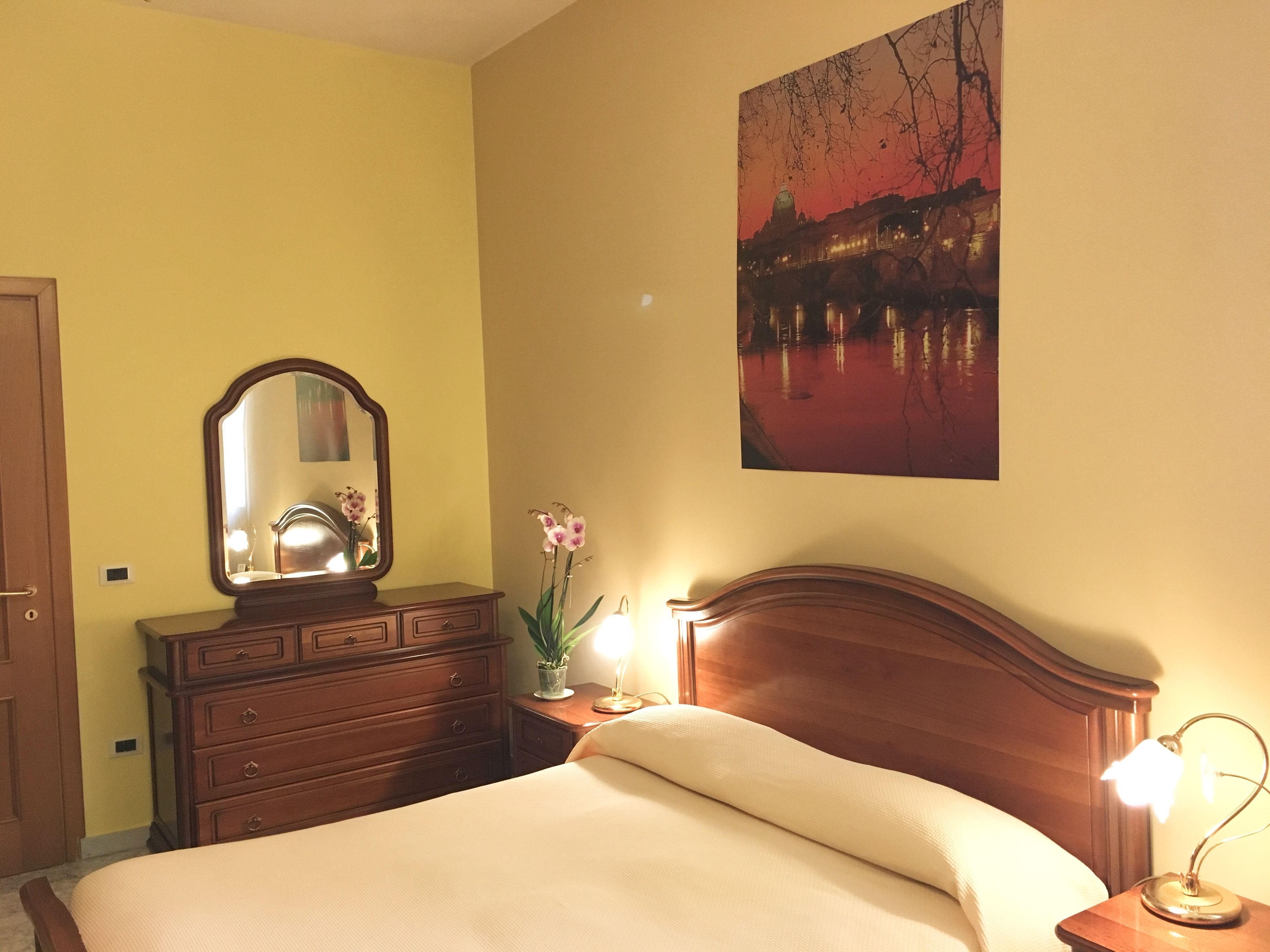 A bedroom with a bed, a dresser with a mirror, a nightstand with a lamp, and a painting of a bridge.