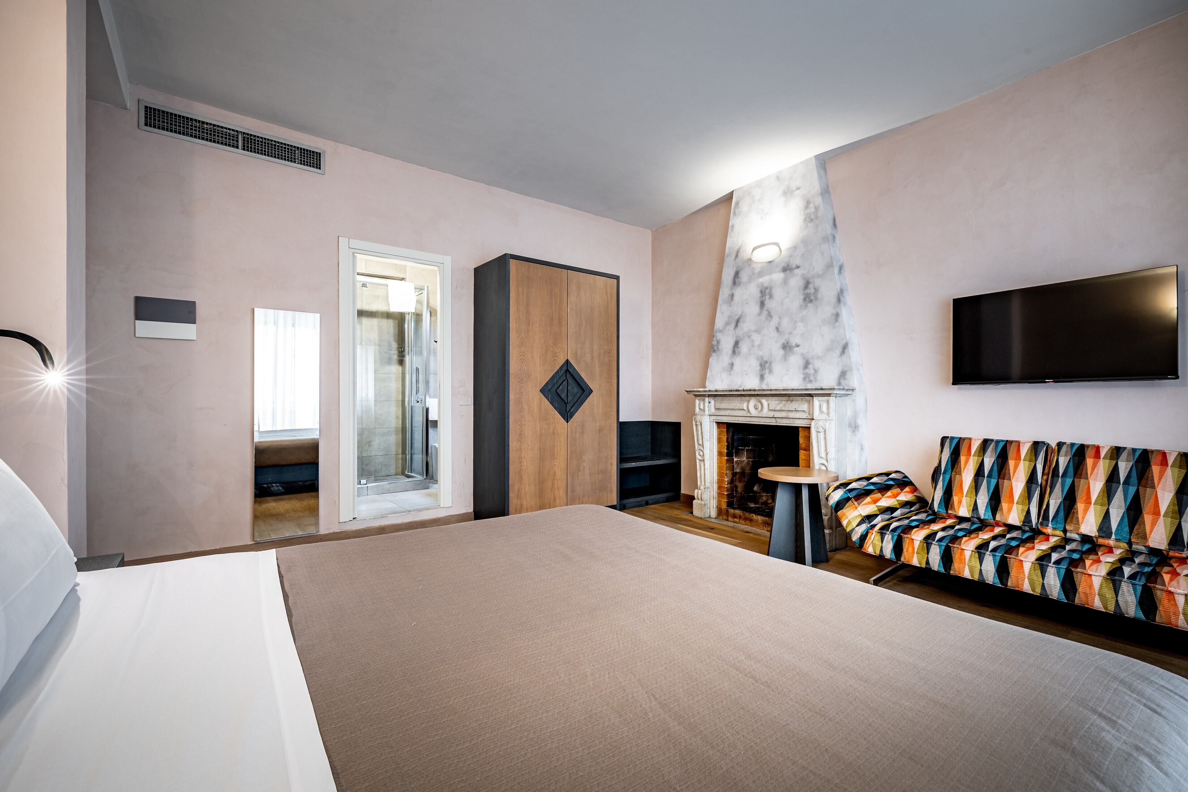 A modern hotel room with a large bed, a fireplace, a TV mounted on the wall, and a colorful sofa.