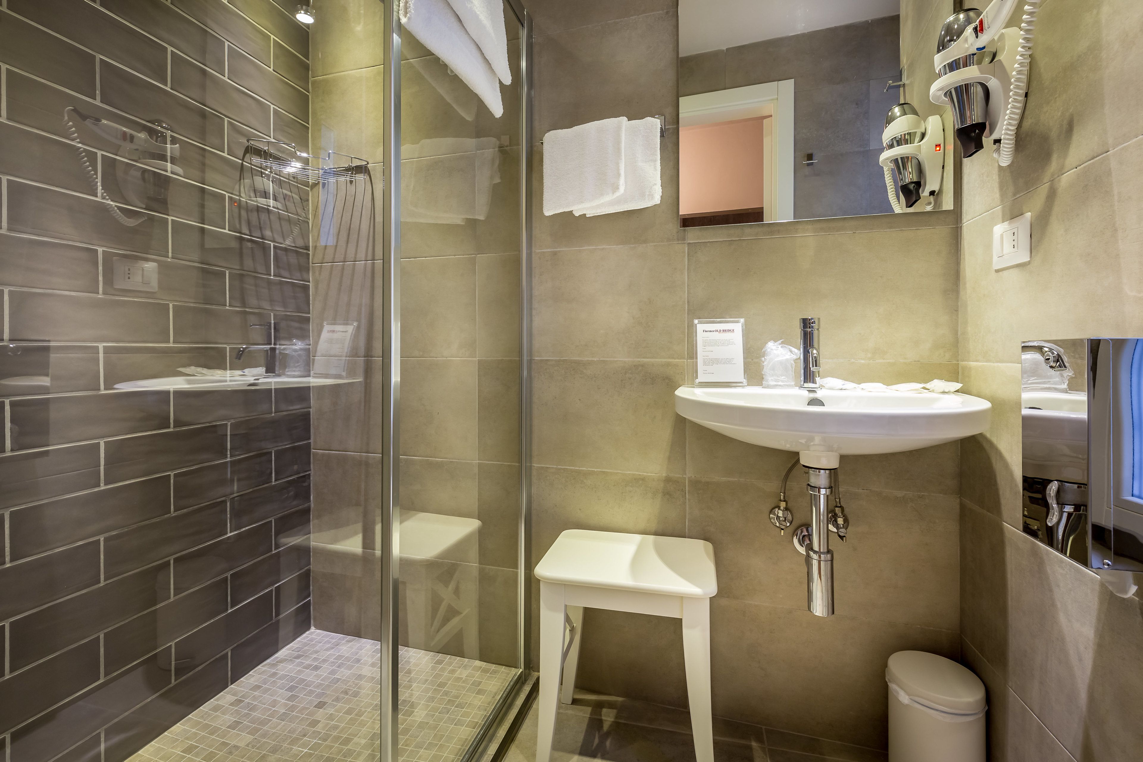 Un baño moderno con ducha acristalada, lavabo blanco y un taburete pequeño.