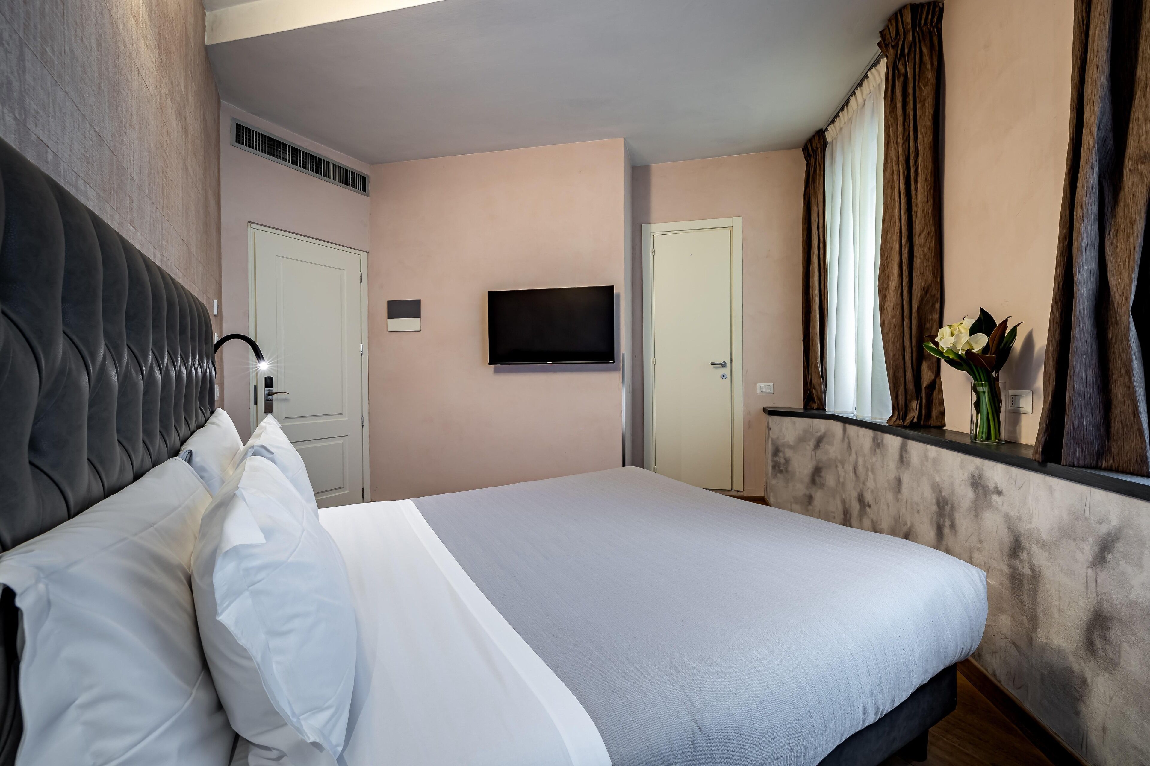 Habitación de hotel con una cama grande, un televisor de pantalla plana montado en la pared, una ventana con cortinas y una pequeña mesa con un florero.