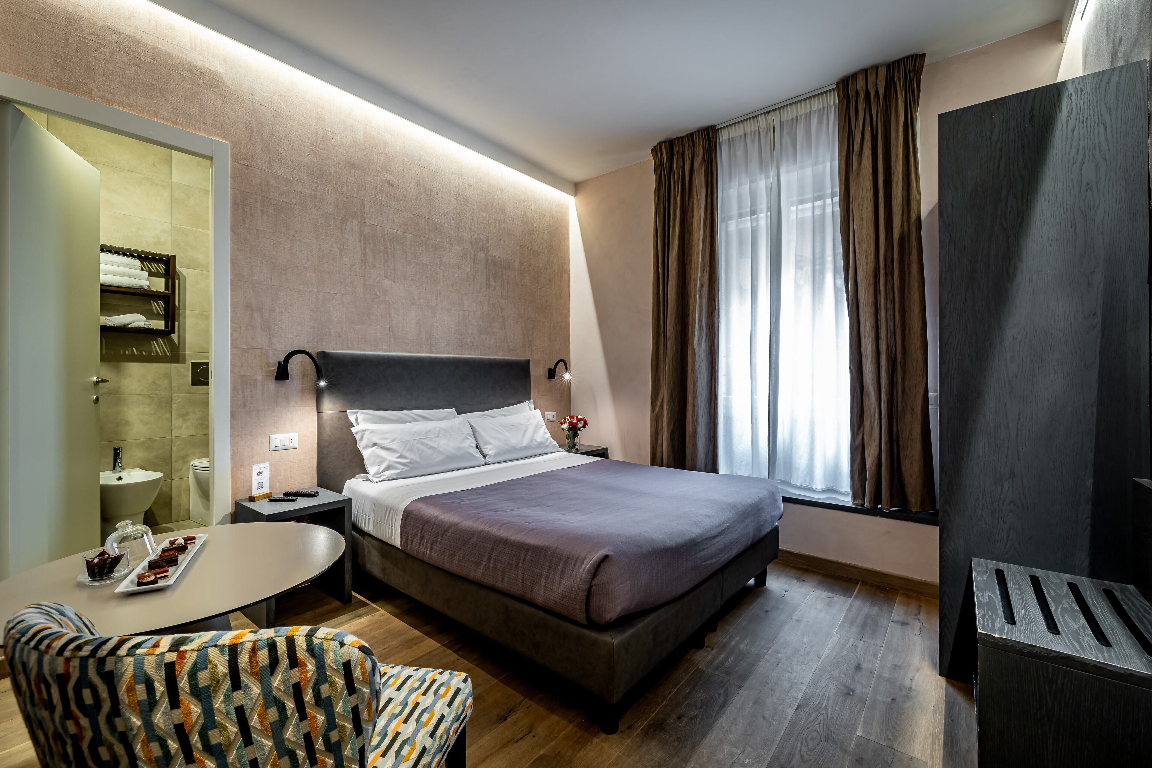 A modern hotel room with a large bed, a bedside table with a lamp, a small round table with a tray of snacks, a patterned armchair, and a large window with curtains.