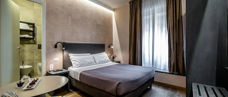 A modern hotel room with a large bed, a bedside table with a lamp, a small round table with a tray of snacks, a patterned armchair, and a large window with curtains.