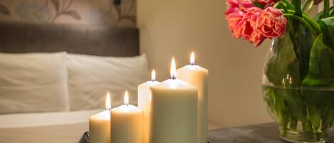 Cuatro velas encendidas sobre una bandeja, un florero con flores rosadas y una cama con ropa blanca de cama al fondo.