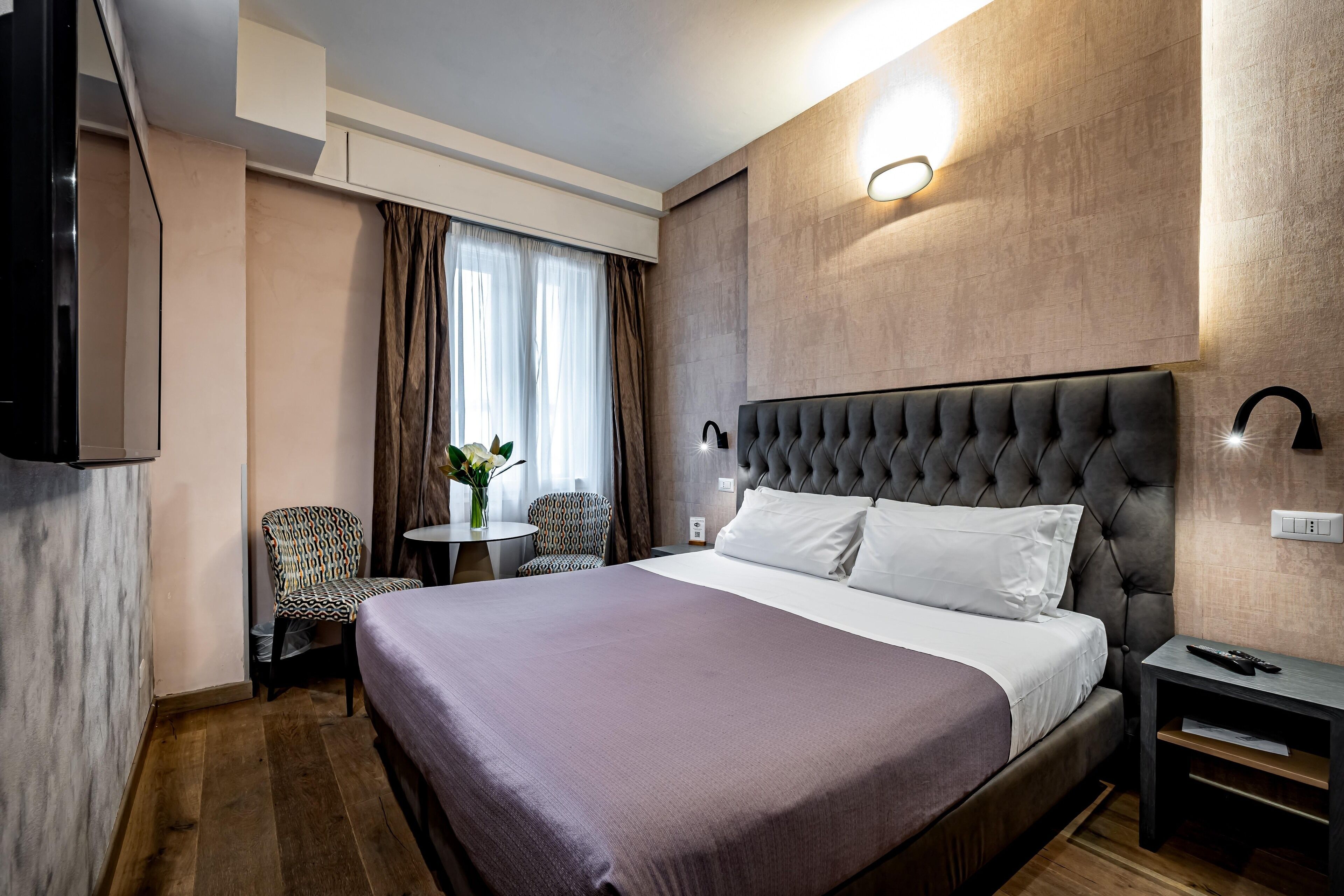 A hotel room with a large bed, a bedside table, a chair, a small table with a vase, and a wall-mounted TV.