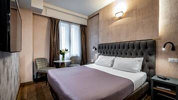 A hotel room with a large bed, a bedside table, a chair, a small table with a vase, and a wall-mounted TV.
