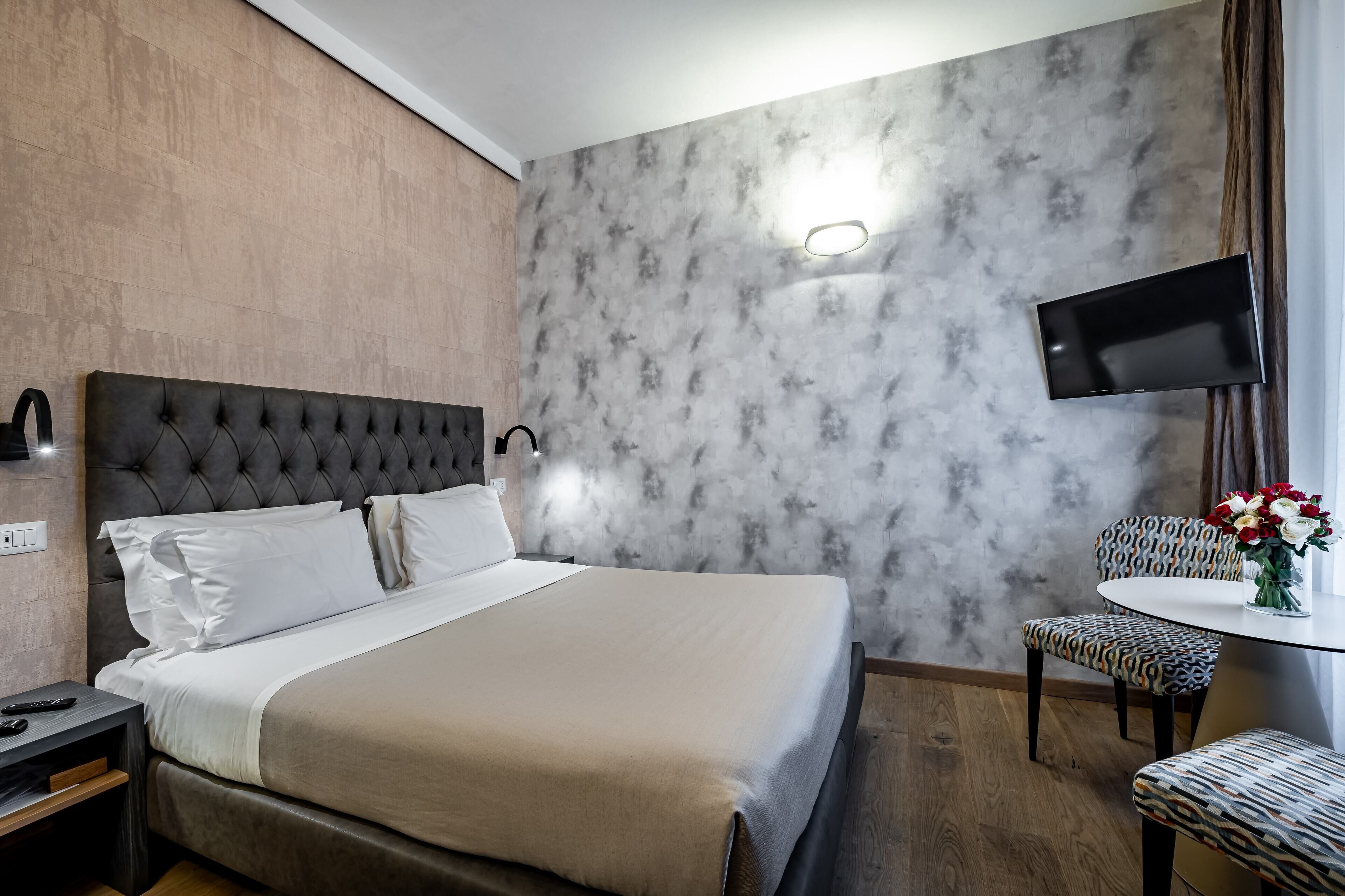 Une chambre d’hôtel avec un grand lit, un téléviseur fixé au mur, une petite table ronde avec une chaise et un vase de fleurs.