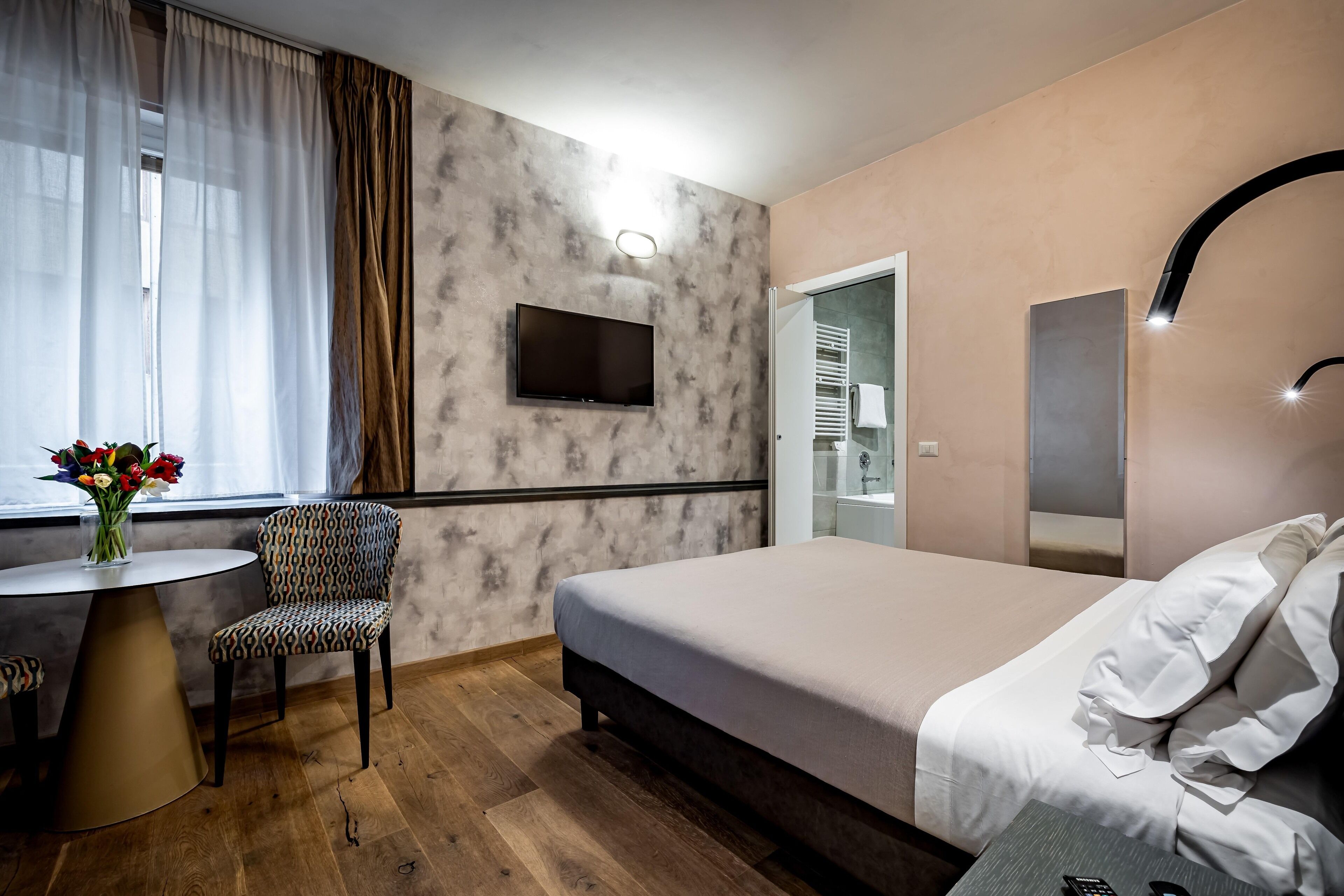 Une chambre d’hôtel avec un lit, un téléviseur fixé au mur, une petite table avec une chaise et une fenêtre avec des rideaux.