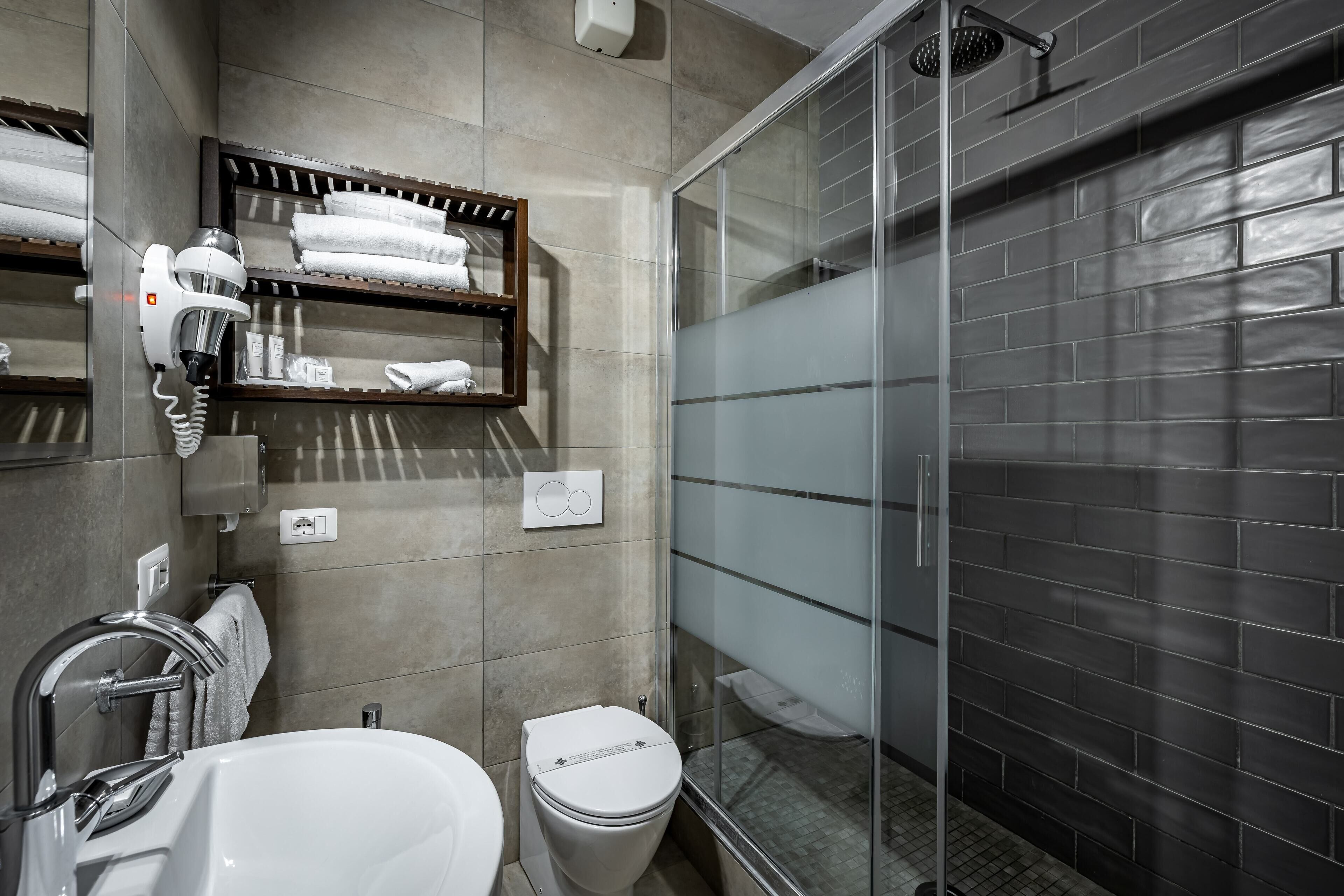 A modern bathroom with a glass shower enclosure, a built-in hairdryer, and neatly arranged towels on shelves.