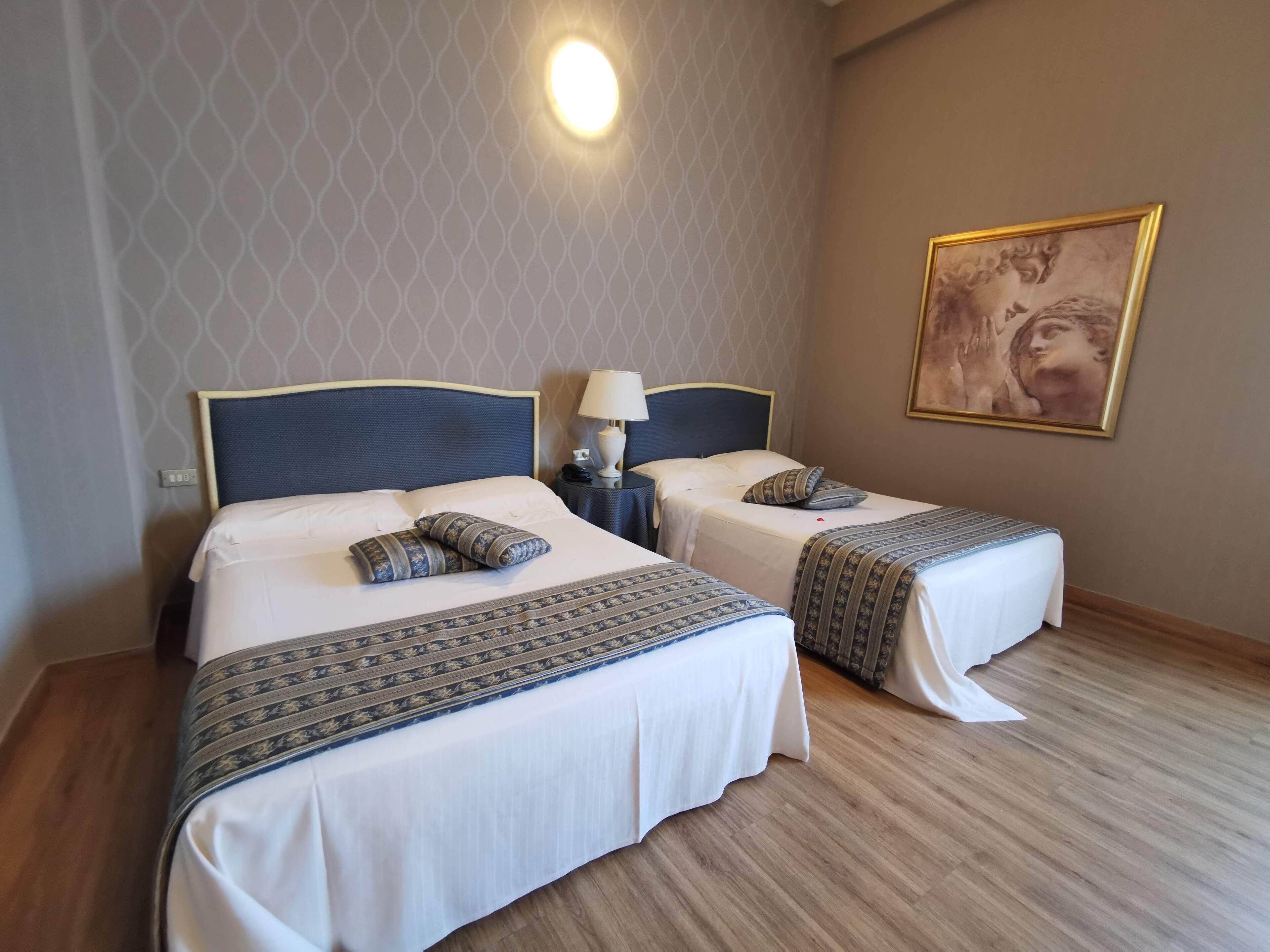 A hotel room with two beds, a nightstand with a lamp, a painting on the wall, and a patterned bedspread.