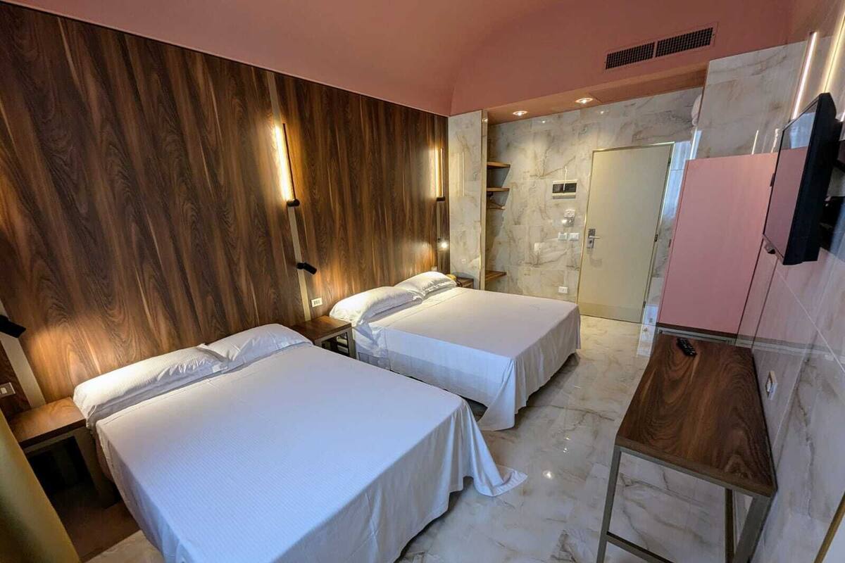 A hotel room with two beds, a wooden headboard, marble flooring, a small desk, and a bathroom with a marble wall and sink.