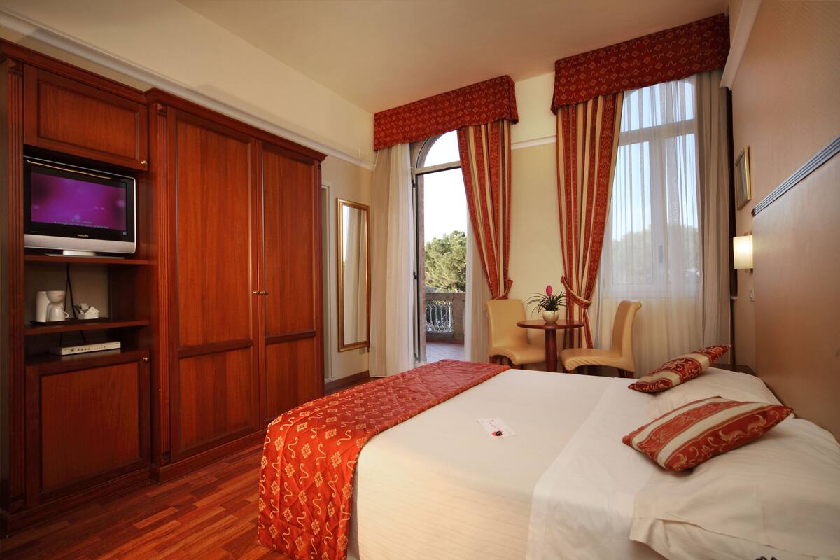 A hotel room with a large bed, a television on a wooden cabinet, a balcony with a chair, and a small table with a plant.