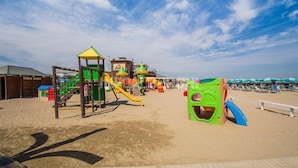 A beachside playground with various play structures, including slides and climbing areas, on a sandy beach with a wooden boardwalk in the foreground. - Hotel River (Rimini)