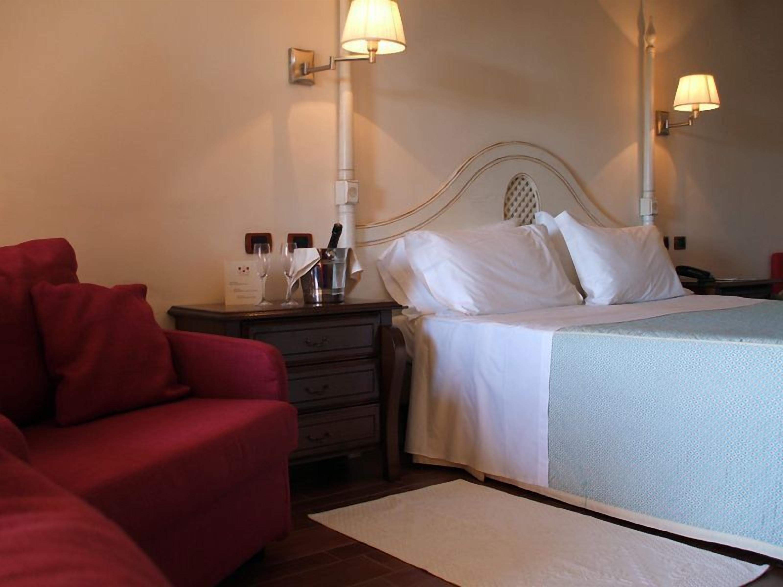 Habitació d'hotel amb un llit, dues làmpades de nit, un sofà vermell, una taula de nit amb una ampolla i vasos, i una taula petita.