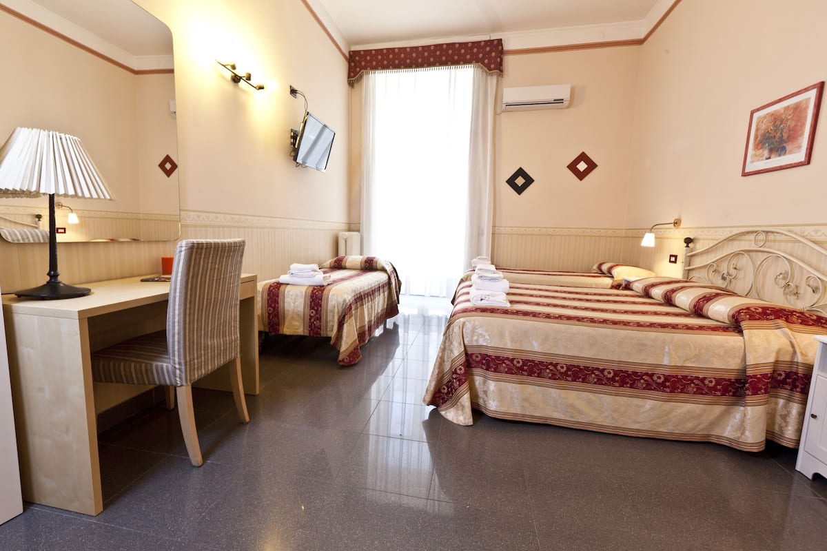Habitación de hotel con escritorio, dos camas individuales, una silla y un ventanal grande.