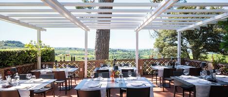 Un área de comedor al aire libre con mesas preparadas para dos personas, rodeada por una estructura de pérgola, con vistas a un paisaje pintoresco.