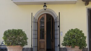 A building with a balcony adorned with purple flowers, a wooden door with an arched entrance, and two potted plants on either side. - La Spiga (Campi Bisenzio)