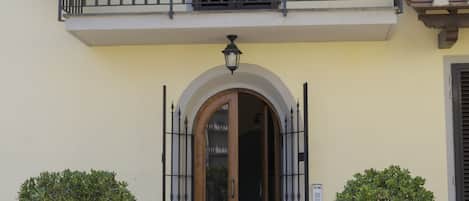 A building with a balcony adorned with purple flowers, a wooden door with an arched entrance, and two potted plants on either side.