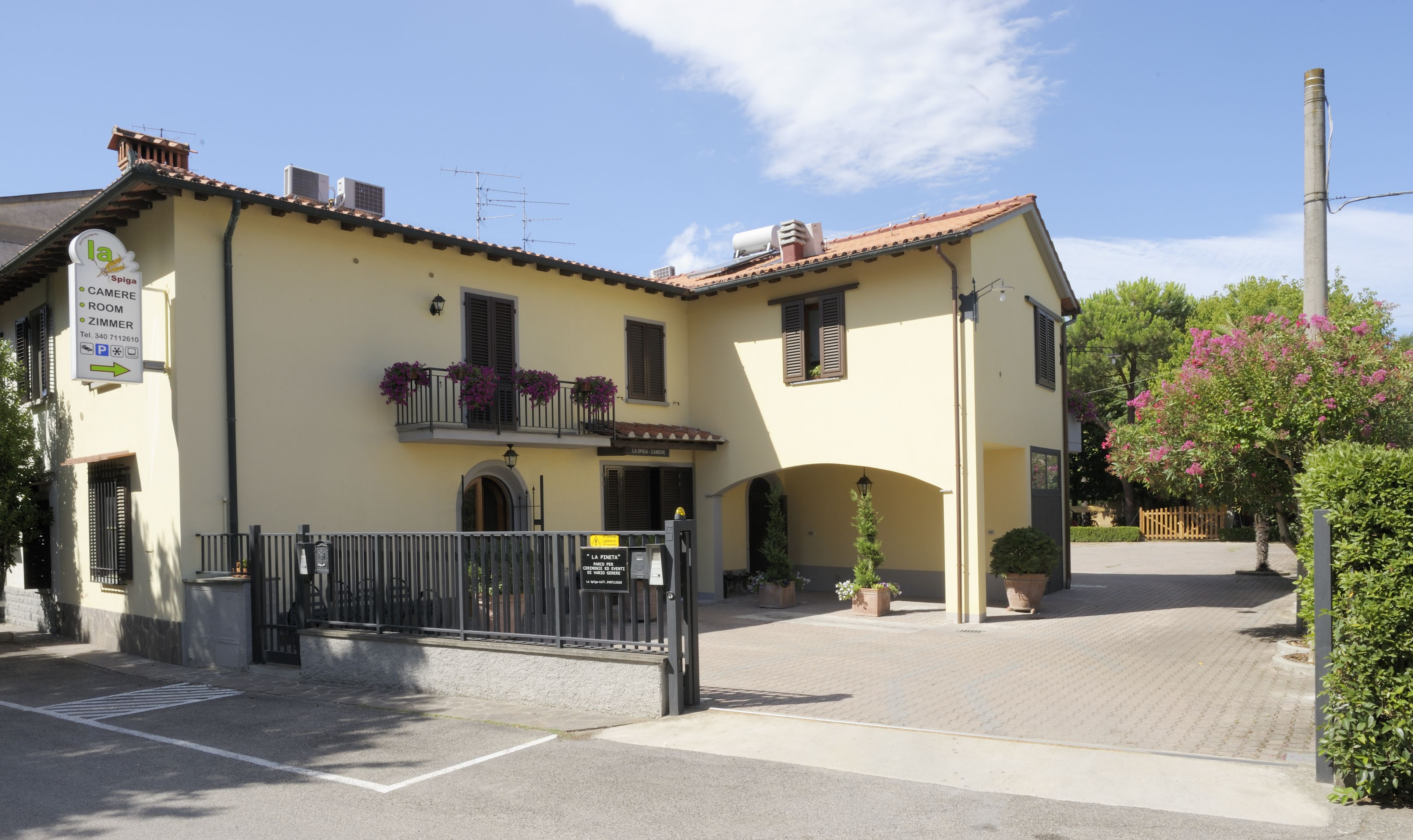 Un edificio de dos pisos con fachada amarilla, un balcón con flores moradas y un cartel que indica disponibilidad de habitaciones y estacionamiento.