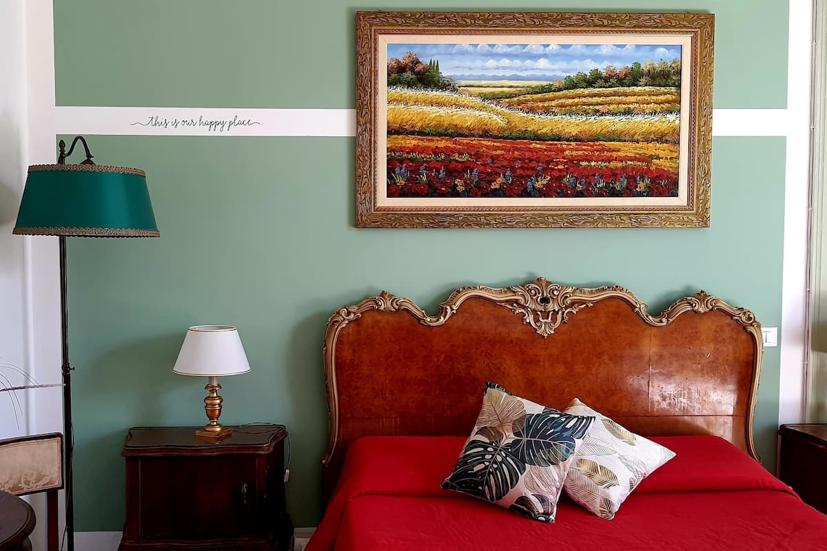 A bedroom with a red bed, a wooden headboard, a framed landscape painting, and a teal floor lamp.