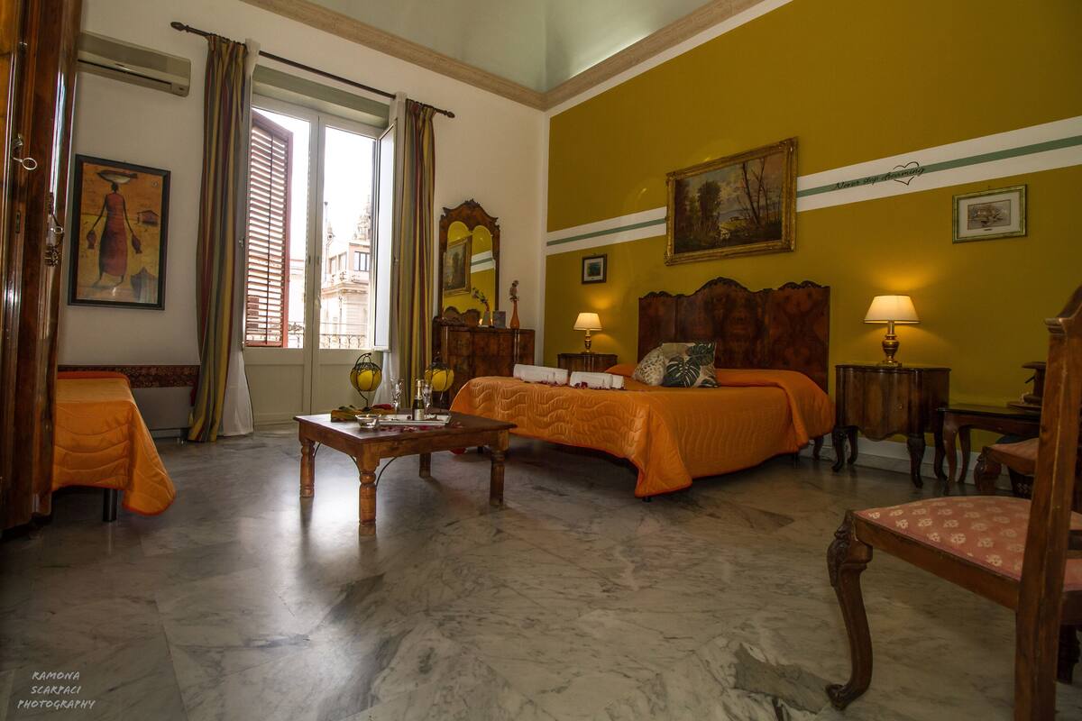 A hotel room with a bed, a desk, a chair, a window with curtains, and a painting on the wall.