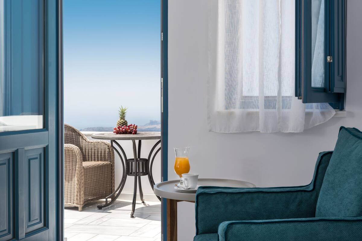 A room with a view of the sea, a wicker chair, a table with a glass of orange juice, and a teal armchair.