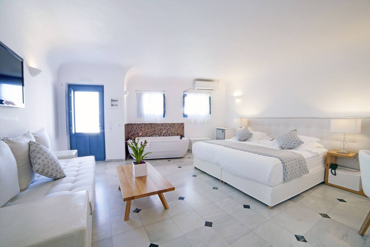A modern, well-lit bedroom with a large bed, a sofa, a small table, and a bathroom with a bathtub.