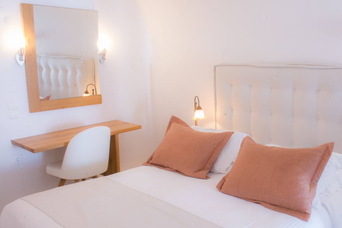 A hotel room with a bed, a desk, a chair, a mirror, and a nightstand with a lamp.