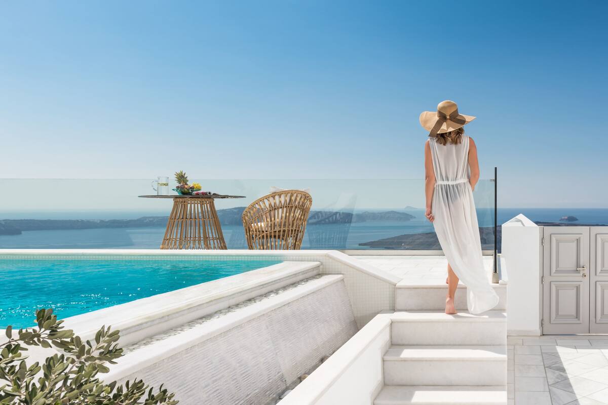 A woman in a white dress and hat standing by a pool with a view of the sea and a distant island.