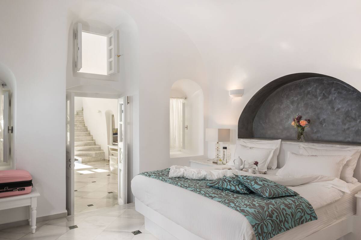 A bedroom with a large bed, a curved headboard, a bedside table with a lamp, and a view of a staircase.