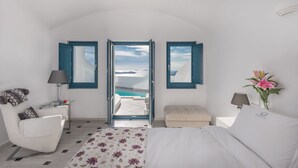 A bedroom with a bed, a chair, a small table, a nightstand, and a view of the sea through open doors. - Anteliz Suites (Santorini)