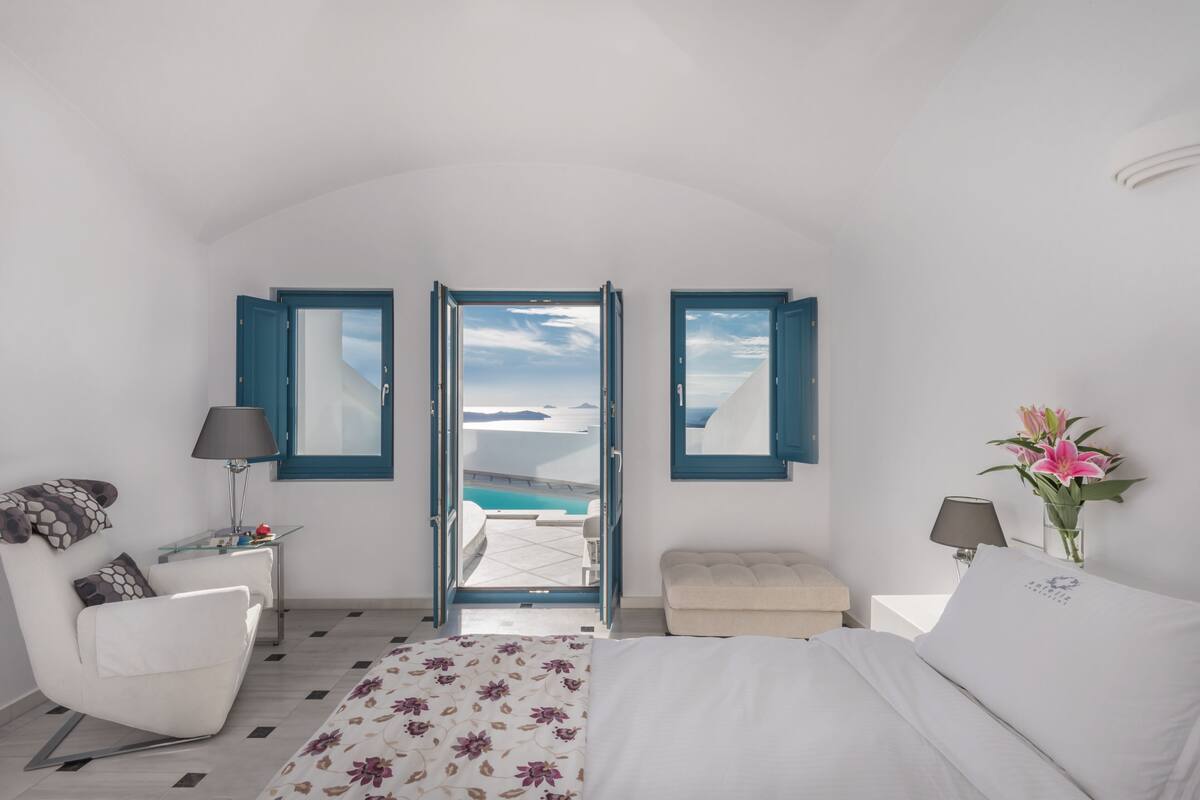 A bedroom with a bed, a chair, a small table, a nightstand, and a view of the sea through open doors.