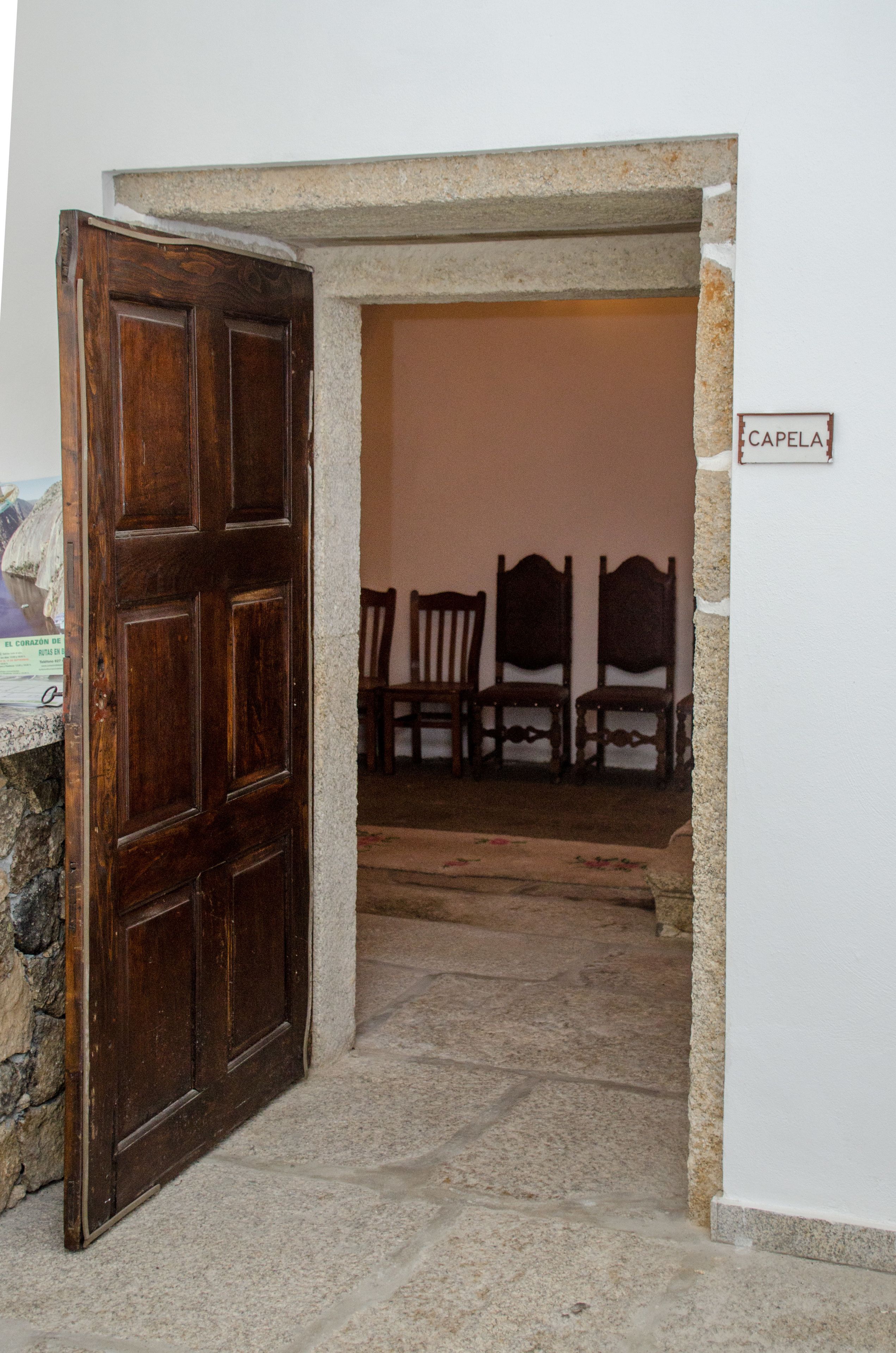 Un vano de puerta que da a una habitación con pared de piedra y puerta de madera. Hay tres sillas de madera en la habitación.