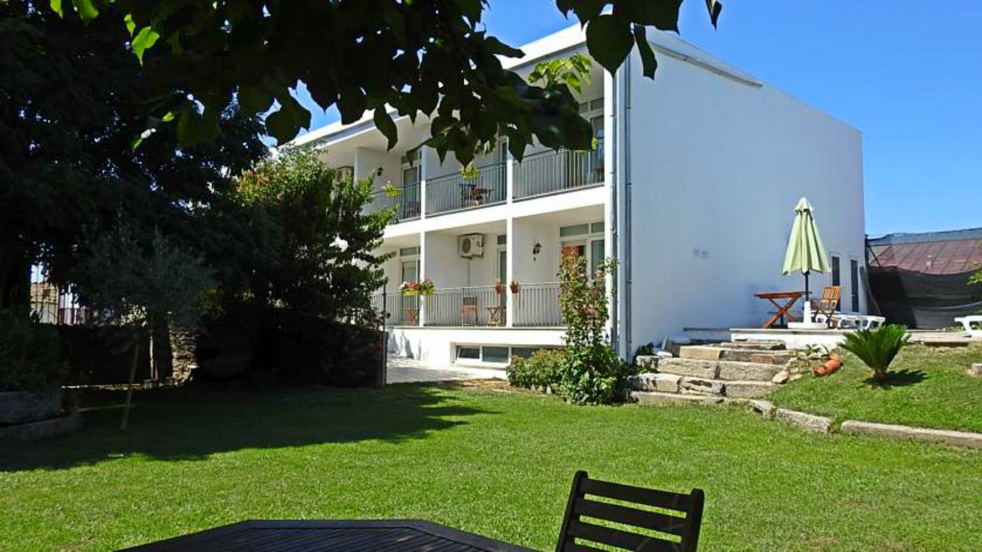 A two-story building with a balcony, a small garden area with a table and chairs, and a green umbrella.