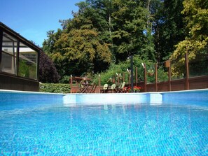 A swimming pool with clear blue water, surrounded by trees and a wooden deck with chairs. - Woodside B&B (Torhout)