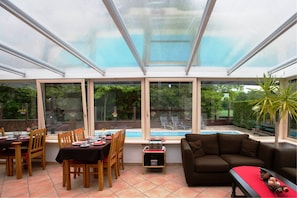 Un solárium con techo de cristal, una zona de comedor con sillas y mesa de madera dispuesta para dos personas, y una piscina visible a través de las ventanas.