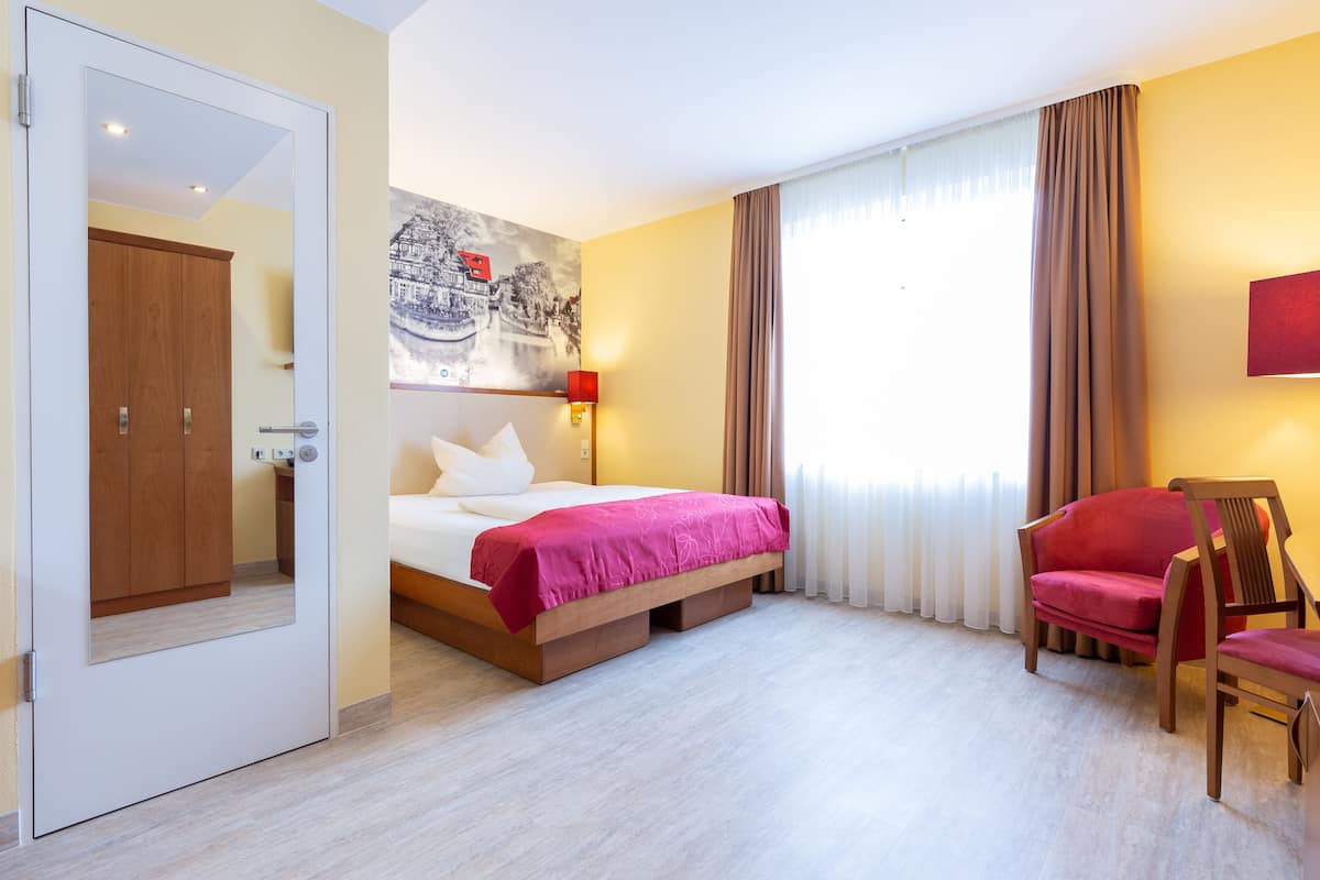 A hotel room with a bed, a chair, a painting on the wall, and a door.