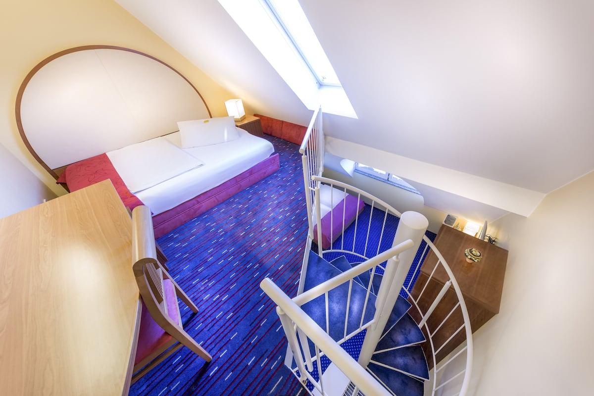 A hotel room with a bed, a desk, a chair, a small table, a nightstand, a lamp, a window, and a staircase.