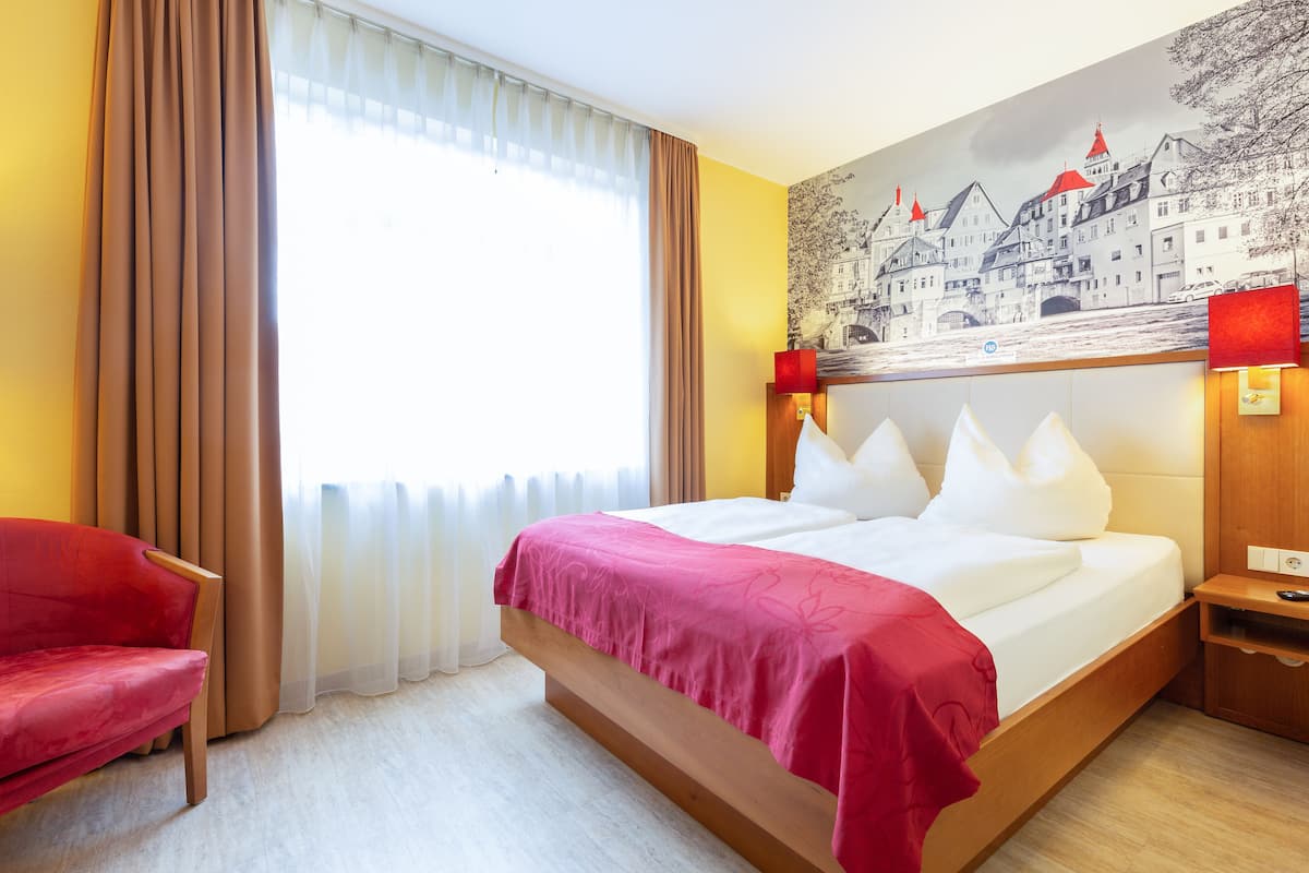 A hotel room with a bed, a red chair, a nightstand, a lamp, and a wall mural.