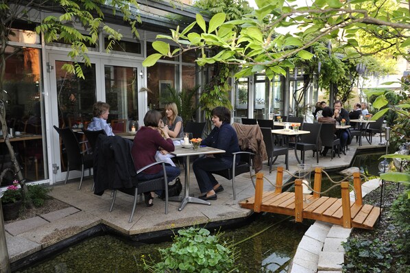 An outdoor cafe with tables and chairs, a small wooden bridge over a water feature, and potted plants.