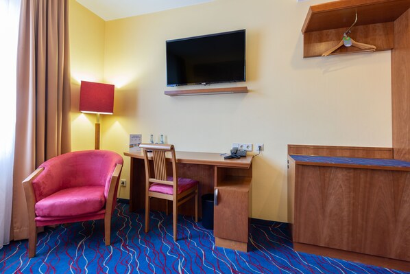 Una habitación de hotel con un escritorio de madera, un televisor montado en la pared, una silla rosa, una lámpara roja y una alfombra estampada.