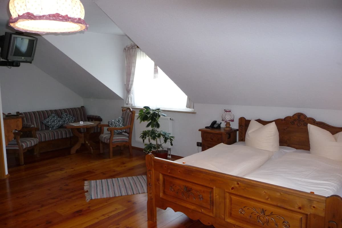A bedroom with a wooden bed, a wooden nightstand, a chair, a small table, a television, and a window with curtains.