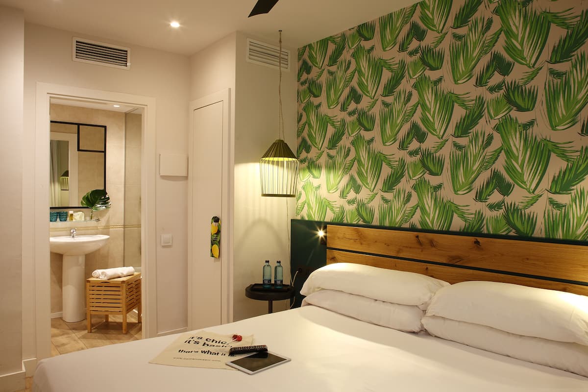 A hotel room with a bed, a bedside table with a lamp, a wall with a leaf pattern, a bathroom with a sink and mirror, and a towel rack.