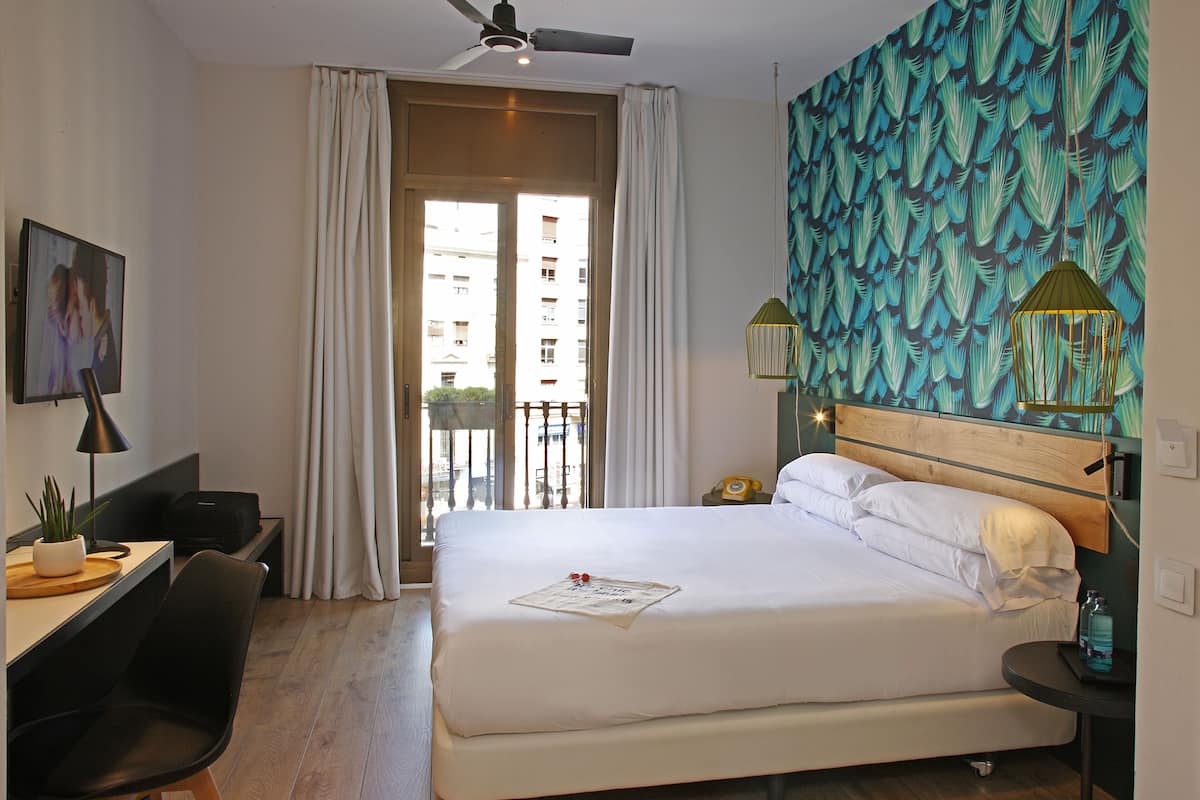 A hotel room with a bed, a desk, a chair, a balcony, and a wall with a leafy pattern.