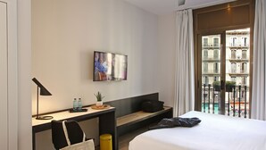 A hotel room with a bed, a desk with a lamp, a TV, a bench, a trash bin, and a view of buildings through the window. - chic&basic Lemon Boutique Hotel (Barcelona)