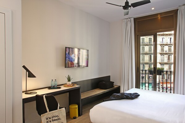 A hotel room with a bed, a desk with a lamp, a TV, a bench, a trash bin, and a view of buildings through the window. - chic&basic Lemon Boutique Hotel (Barcelona)