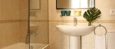 A bathroom with a bathtub, sink, and toilet. There is a yellow trash bin and a green plant on the sink.