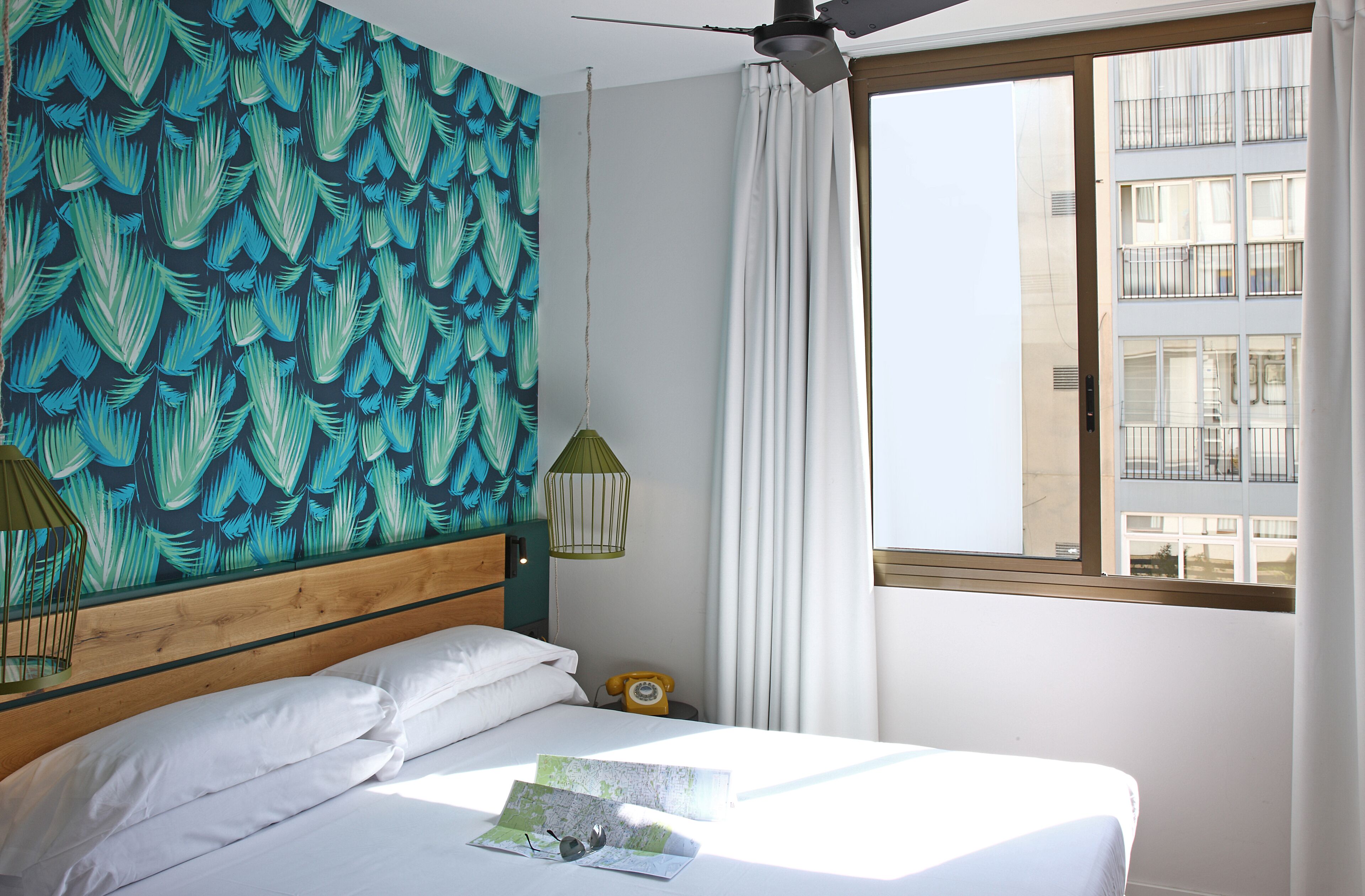 A hotel room with a bed, a ceiling fan, a window with curtains, a wall with a leaf pattern, and a nightstand with a lamp and a telephone.